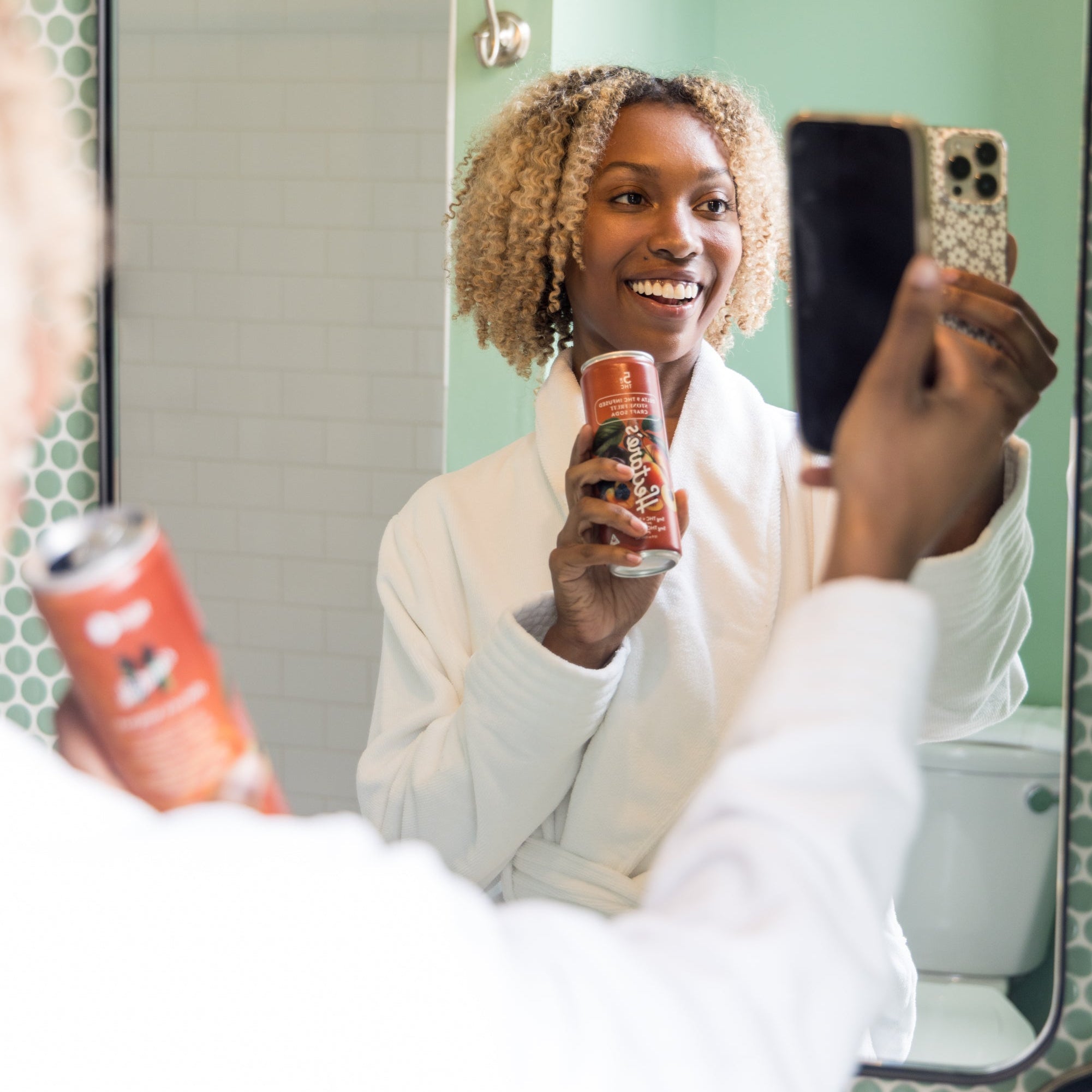Woman in a bathrobe holding a phone and a can of Hectare's Stone Fruit Delta 9 Craft Soda in front of a mirror.