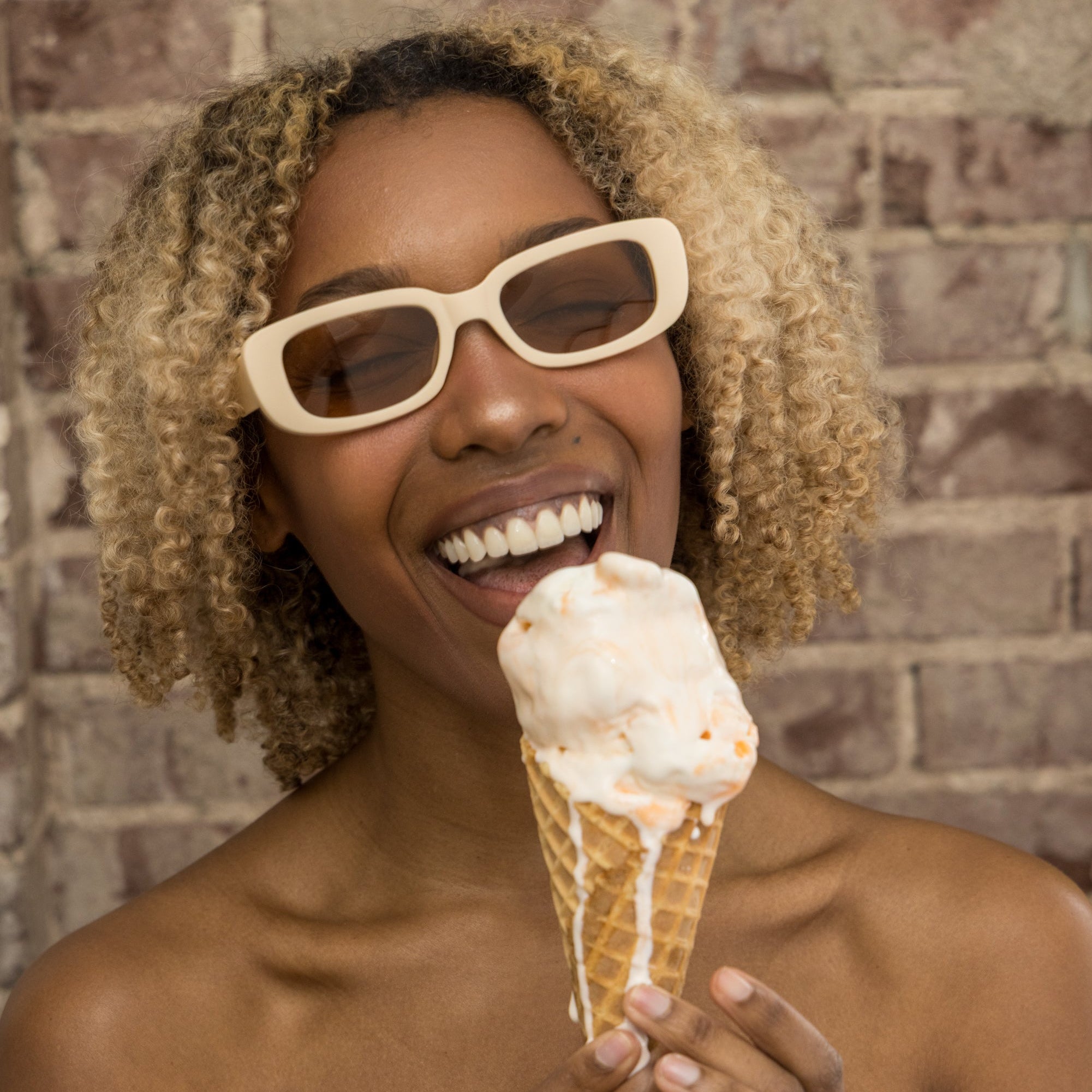 Woman wearing sunglasses and a white dress holding an ice cream cone against a brick wall.